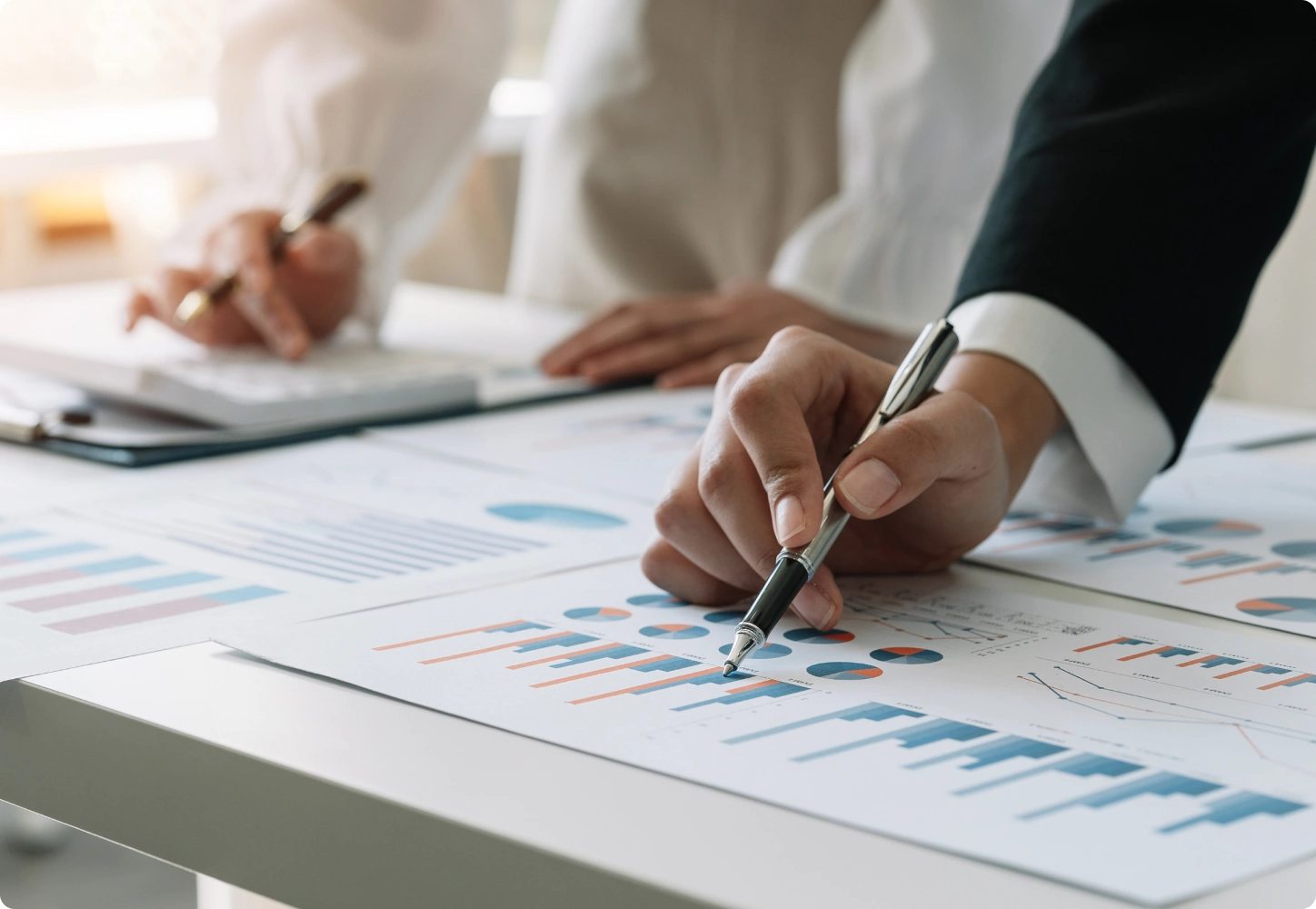 Two people are analyzing charts and graphs on printed documents, with one hand pointing at a bar graph using a pen.